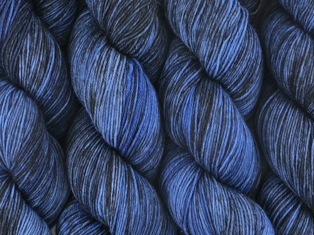 A close up of variegated yarn of black and charcoal with overtones of denim, chambray and navy coloured skeins of superwash merino and nylon 4ply fingering sock yarn (Moody Blues on Tough Stocking)